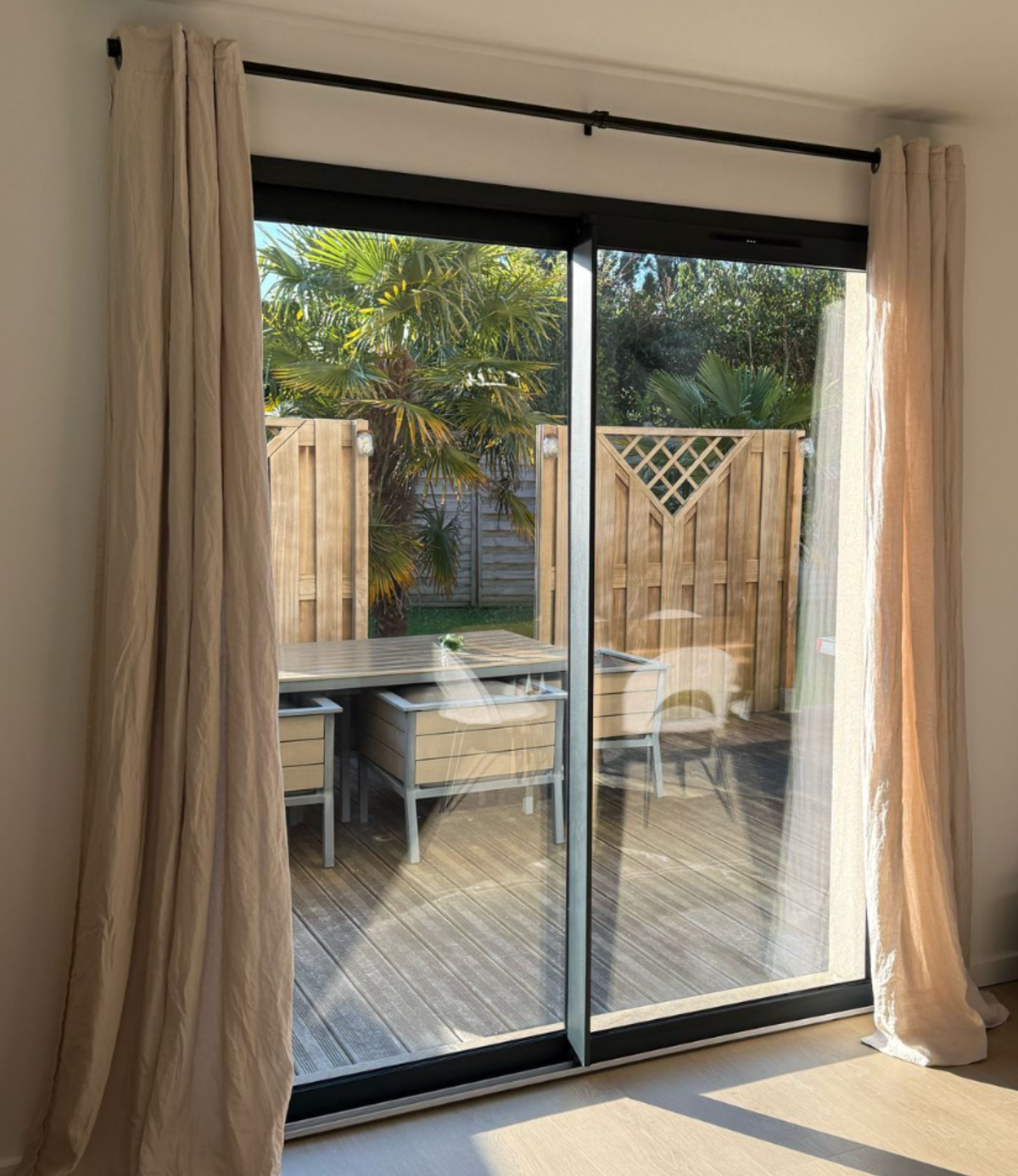 Pièce lumineuse, baie coulissante alu noir à rideaux beige ouvrant sur terrasse bois et salon de jardin