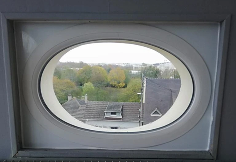 Fenêtre fixe ovale blanche dans cadre gris, radiateur vertical gris, toits et arbres automne