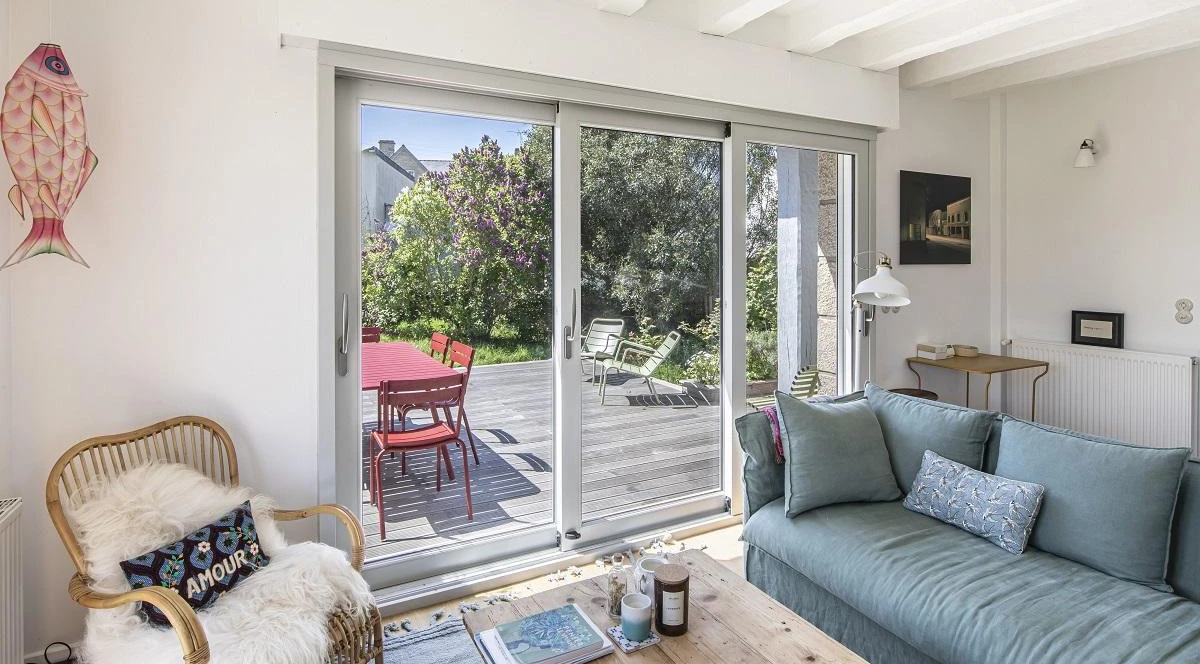 Baie coulissante grise trois vantaux, salon bleu, fauteuil osier et terrasse bois fleurie