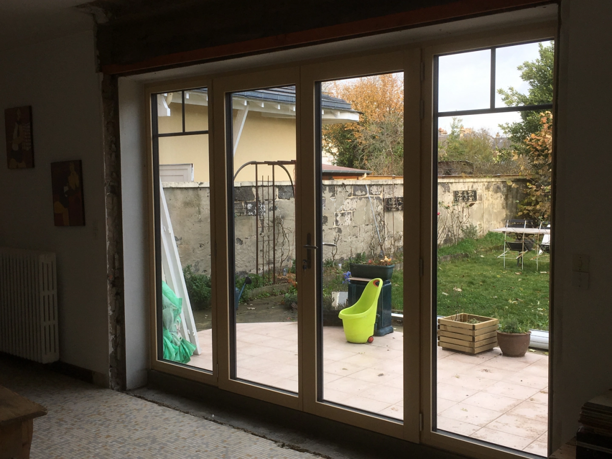 Quatre portes-fenêtres crème à petits carreaux, mur brut chantier, vue terrasse et jardin