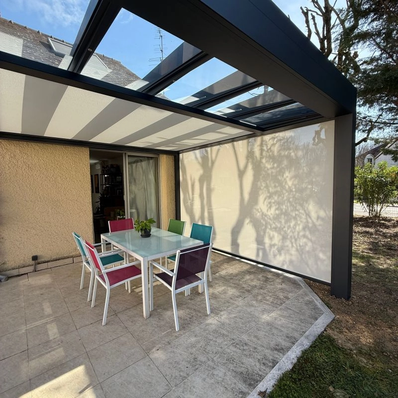 Pergola gris foncé, toiture toile rayée et vitrage, table blanche et chaises colorées