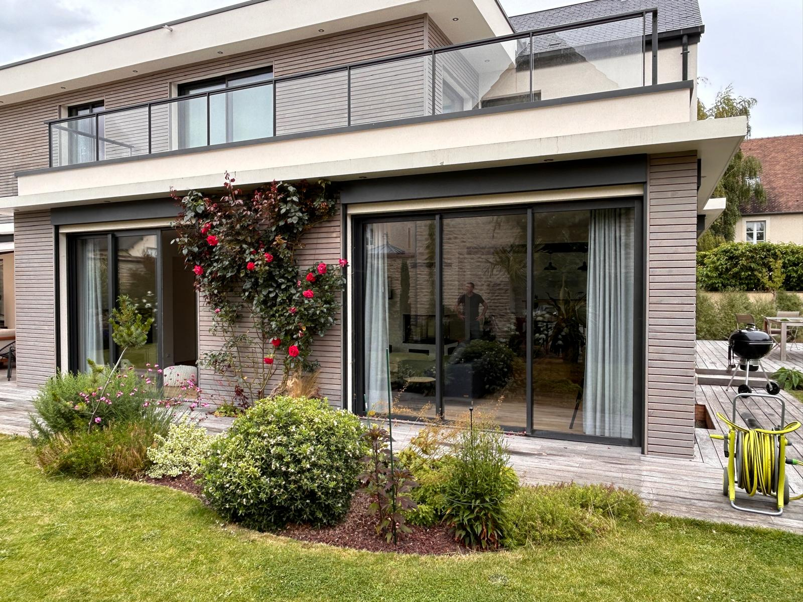Façade moderne avec grandes baies coulissantes alu noir ouvrant sur une terrasse bois et pelouse
