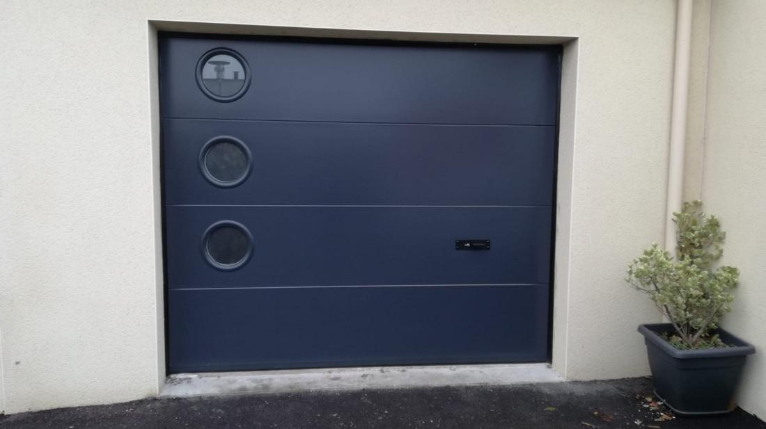 Porte garage sectionnelle bleu anthracite, trois hublots à gauche, poignée noire, pavés