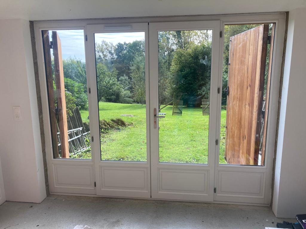 Grande porte-fenêtre blanche à quatre vantaux avec soubassements, vue sur jardin et volets bois ouverts