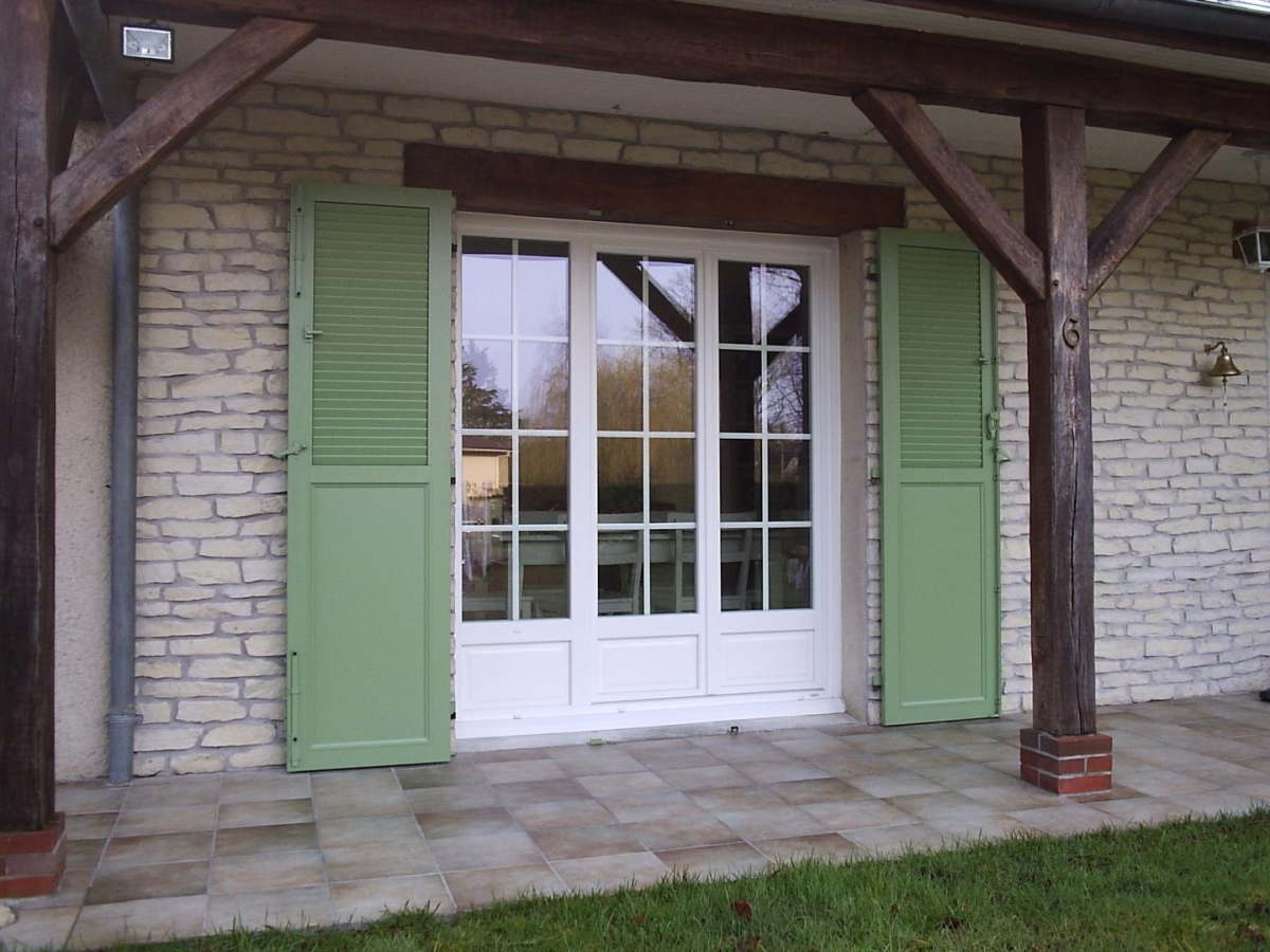 Porte-fenêtre blanche petits bois, volets vert pâle persiennes, mur pierre, terrasse