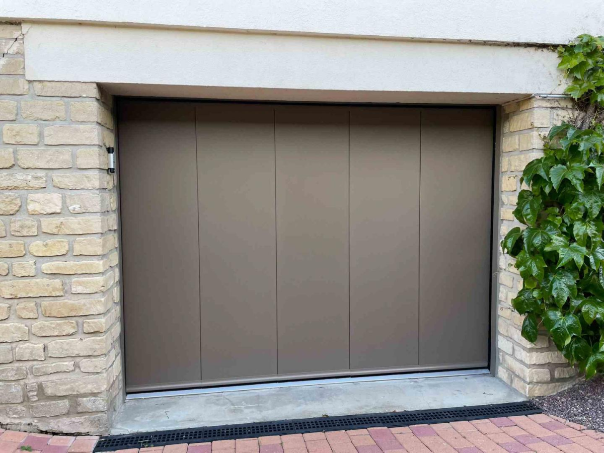 Porte garage latérale bronze, cinq panneaux verticaux, mur pierres claires et lierre