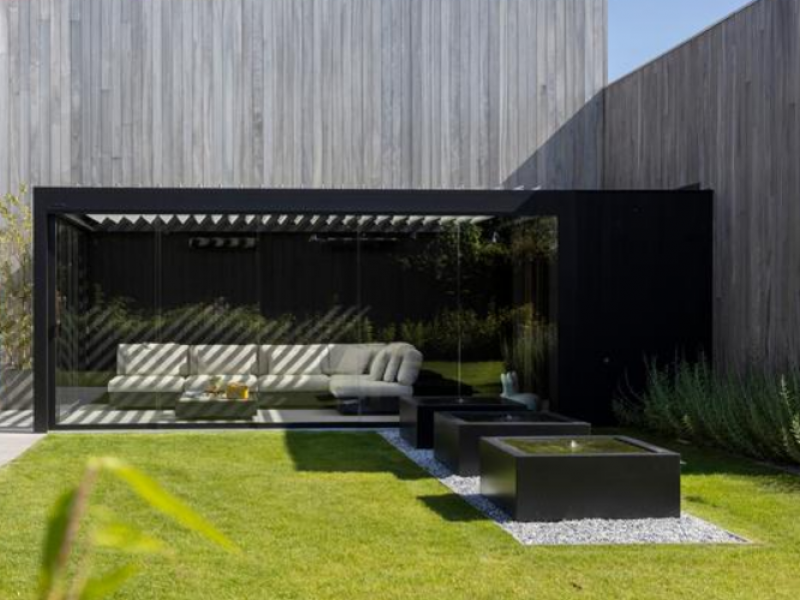 Pergola bioclimatique noire vitrée, canapé clair, pelouse et trois fontaines carrées