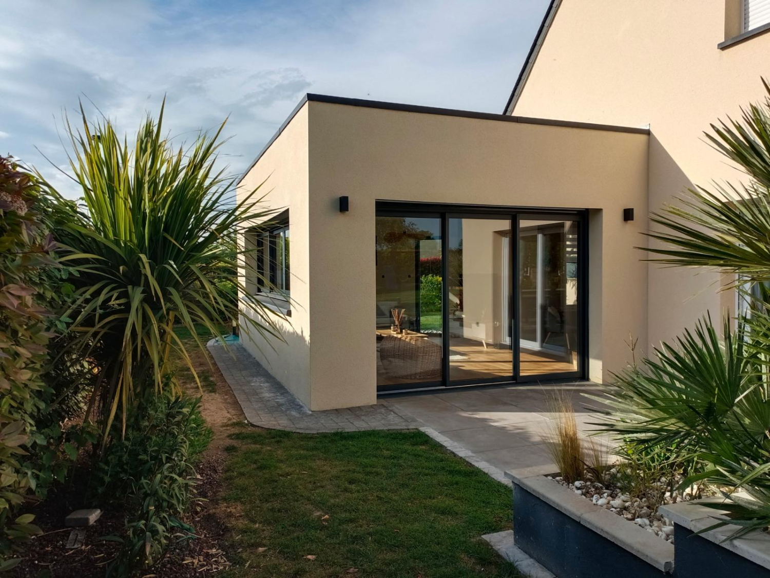 Extension cubique enduit beige, grandes baies gris anthracite, terrasse dalles et plantes