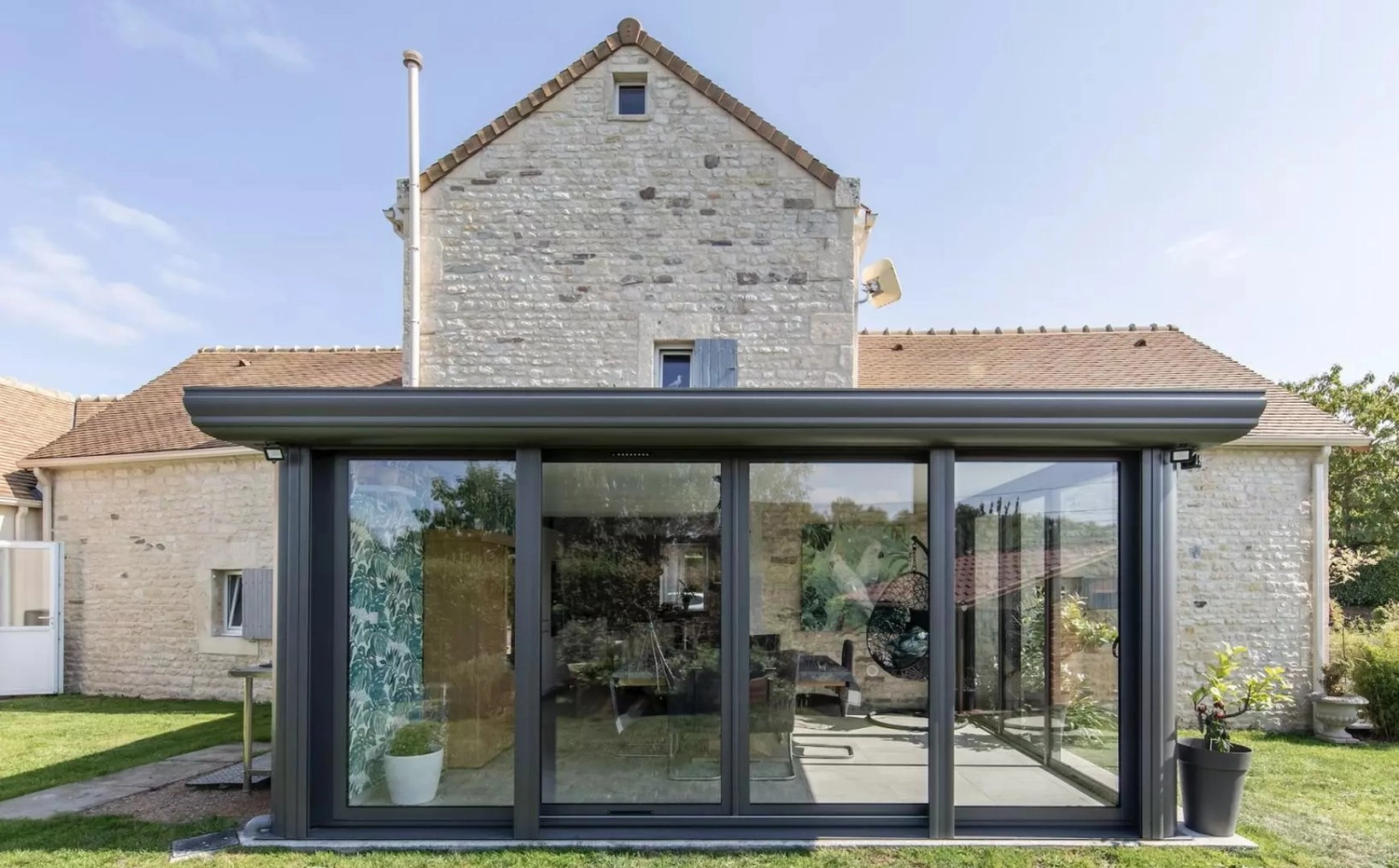 Extension vitrée gris anthracite sur maison pierre, pelouse verte et grand pot gris