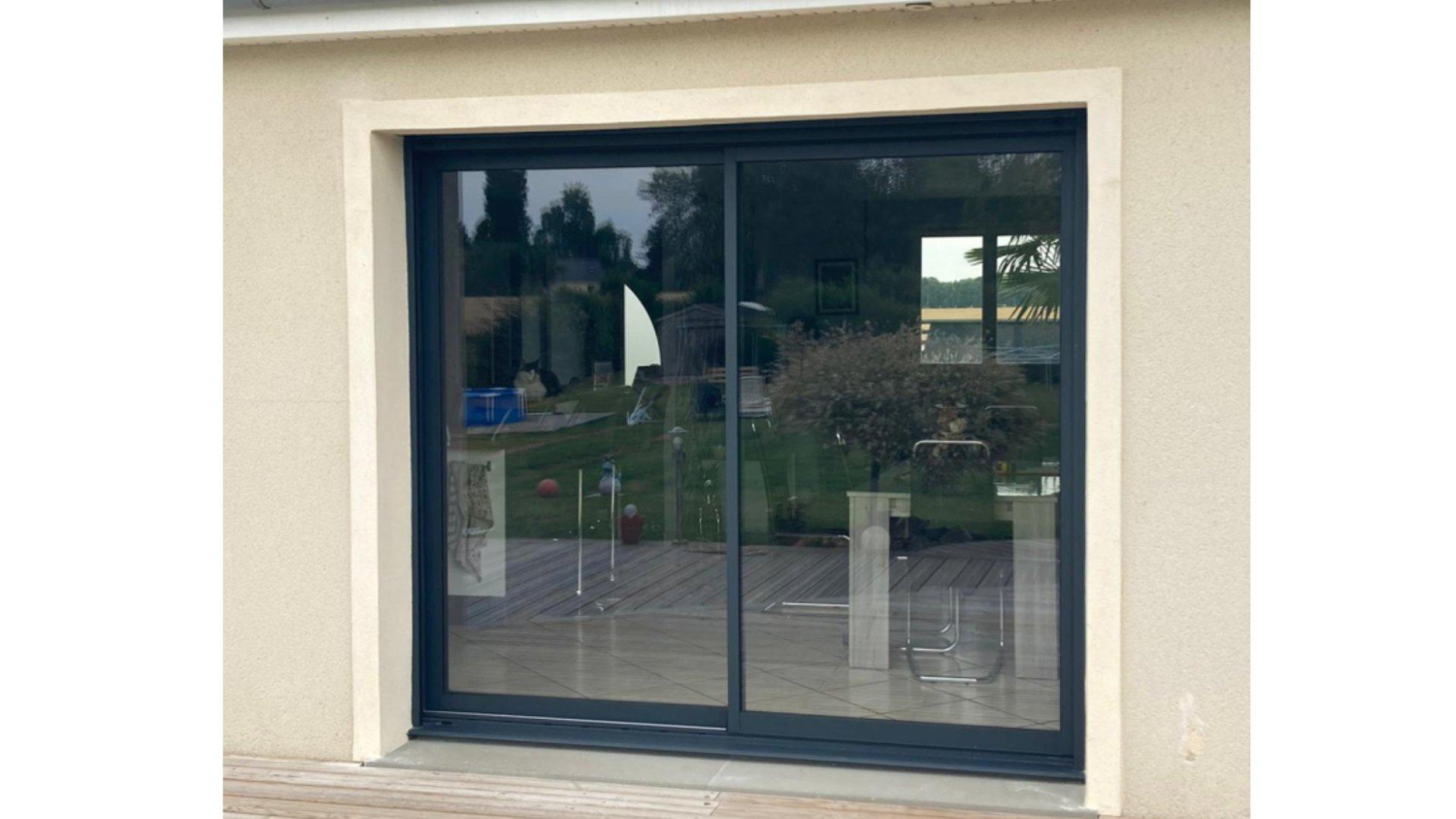 Baie coulissante gris anthracite sur enduit beige, reflets jardin et terrasse bois