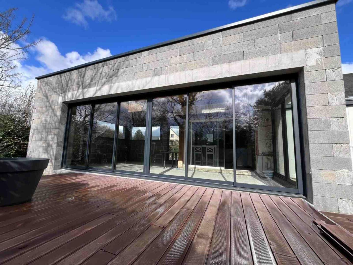 Baie coulissante alu anthracite quatre vantaux sur terrasse bois et mur en pierres grises