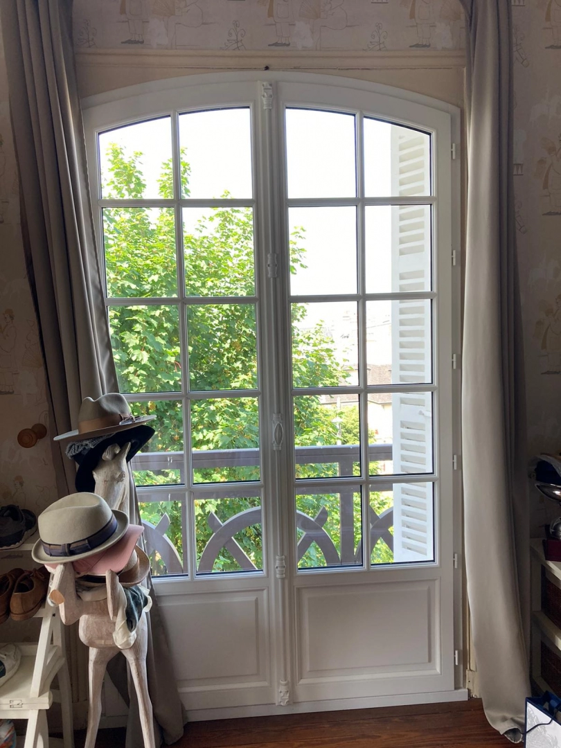 Porte-fenêtre blanche cintrée à petits carreaux, rideaux beige et sculpture cheval chapeaux