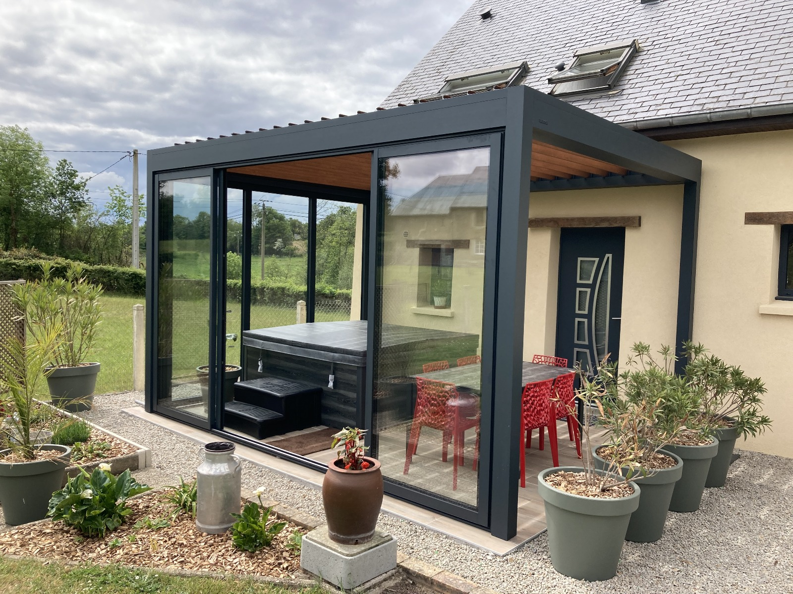 Pergola alu anthracite à parois vitrées abritant un spa bâché et un coin repas aux chaises rouges
