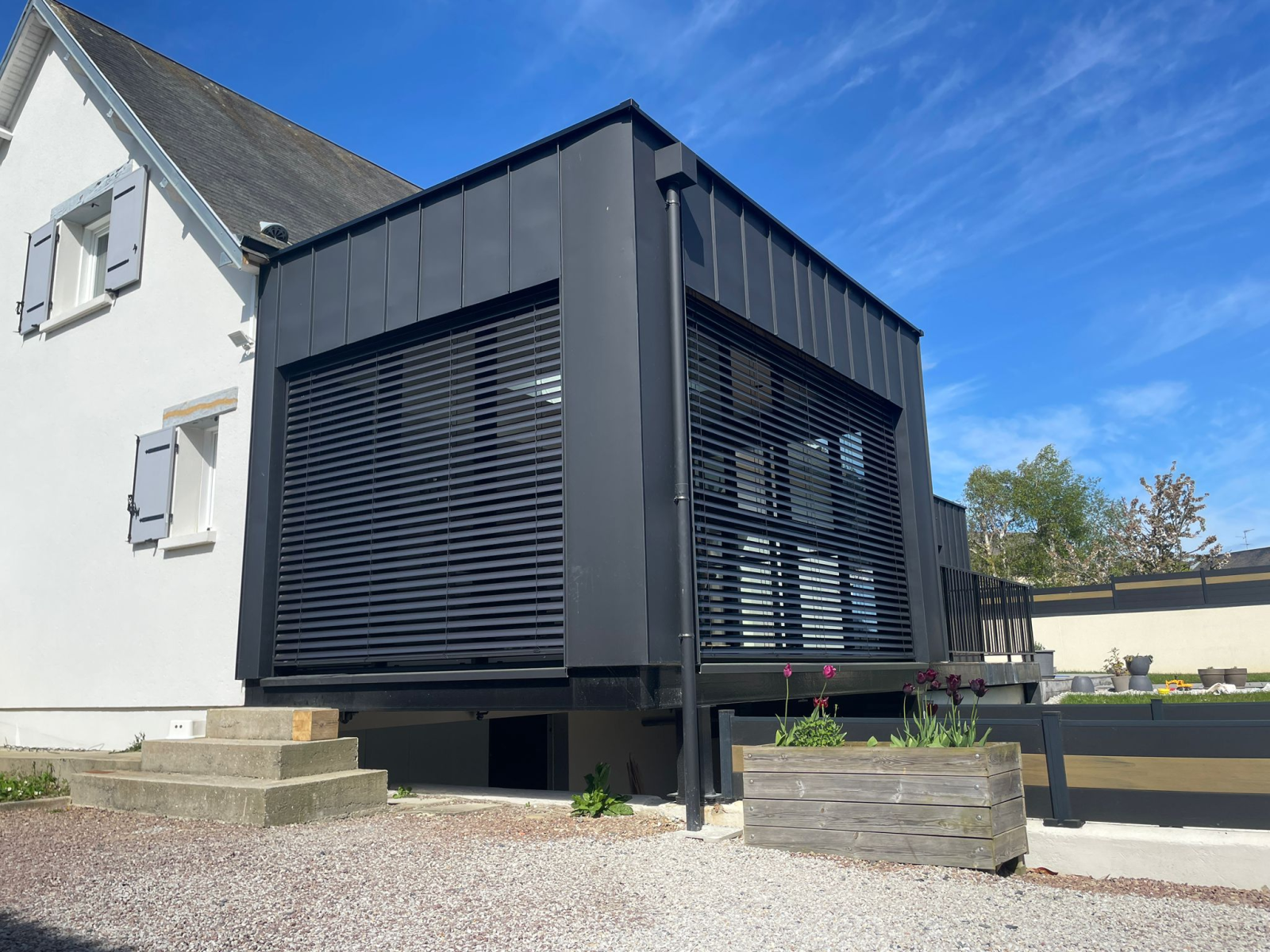 Extension métal noir avec brise-soleil orientables noirs, adossée à maison blanche volets gris