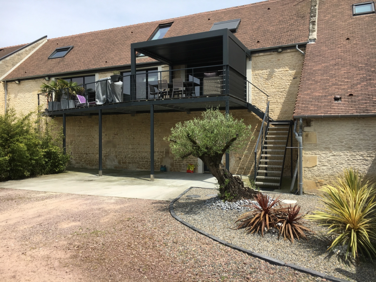 Terrasse sur pilotis noire avec pergola sur facade en pierre et olivier au pied