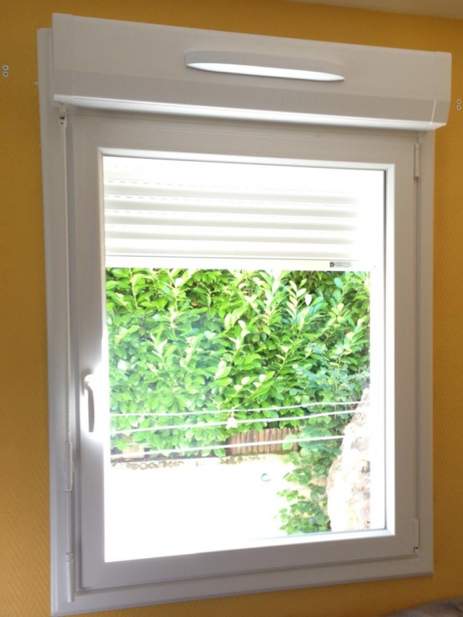 Fenêtre PVC blanche, volet roulant solaire intégré partiellement baissé, haie verte