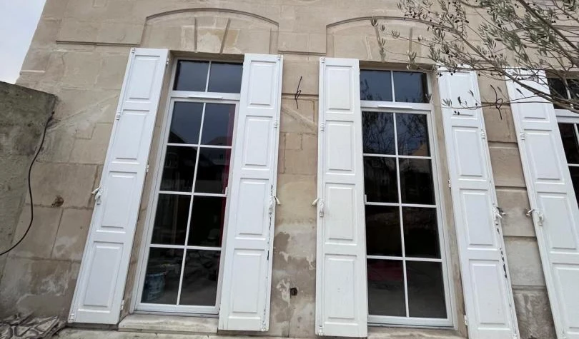 Fenêtres hautes blanches à petits carreaux, volets blancs ouverts sur mur pierre