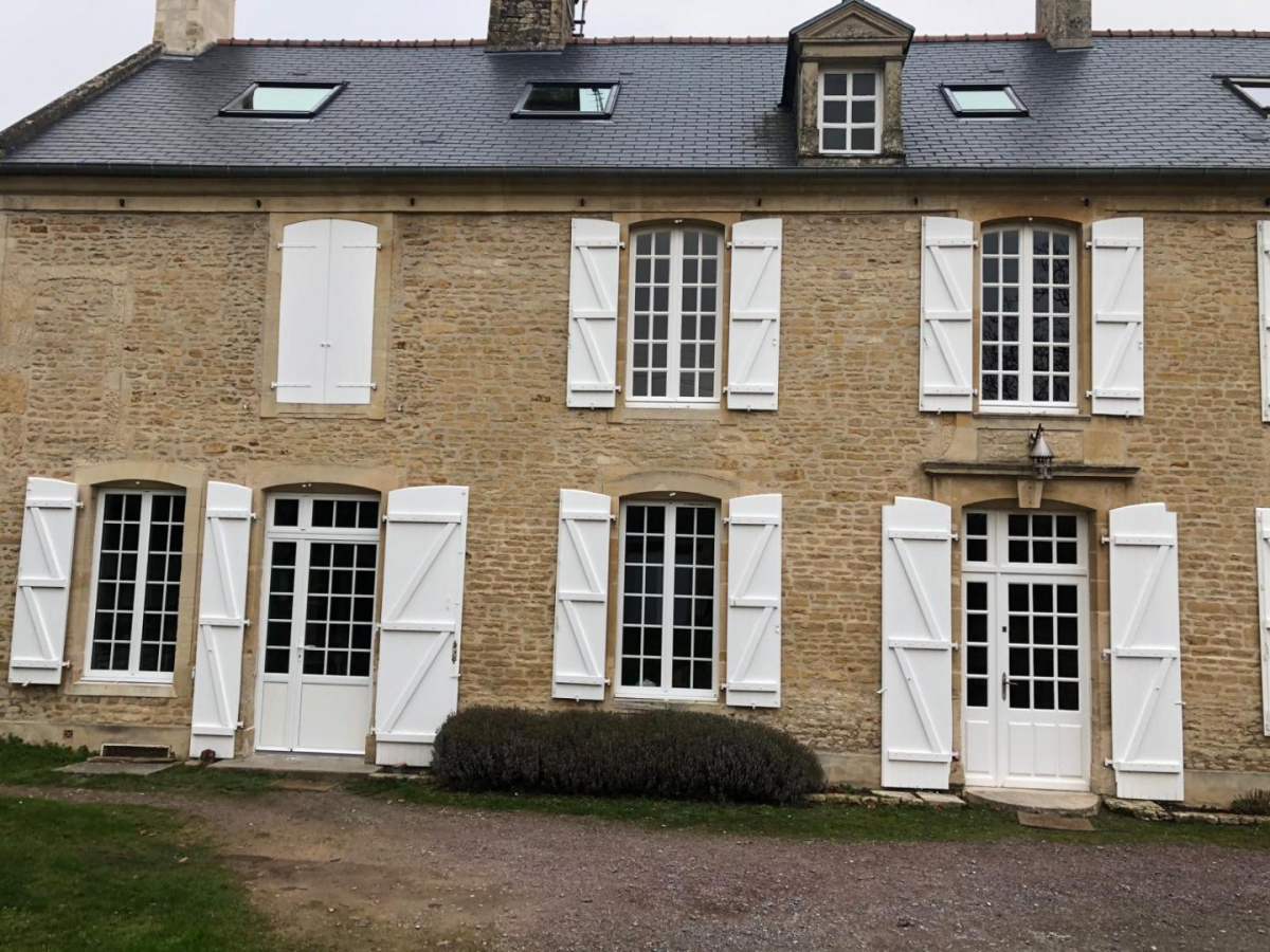 Façade pierre claire, fenêtres et portes blanches à petits carreaux, volets blancs ouverts