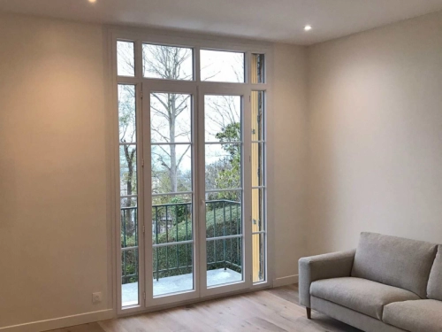 Porte-fenêtre blanche à petits bois, parquet clair, canapé gris et vue arbres depuis balcon