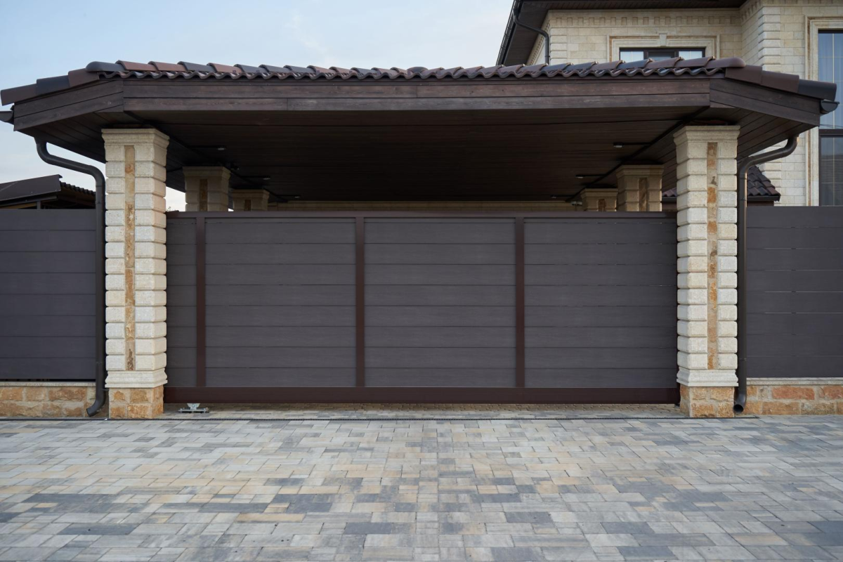 Portail coulissant brun lames horizontales sous carport tuiles entre piliers pierre