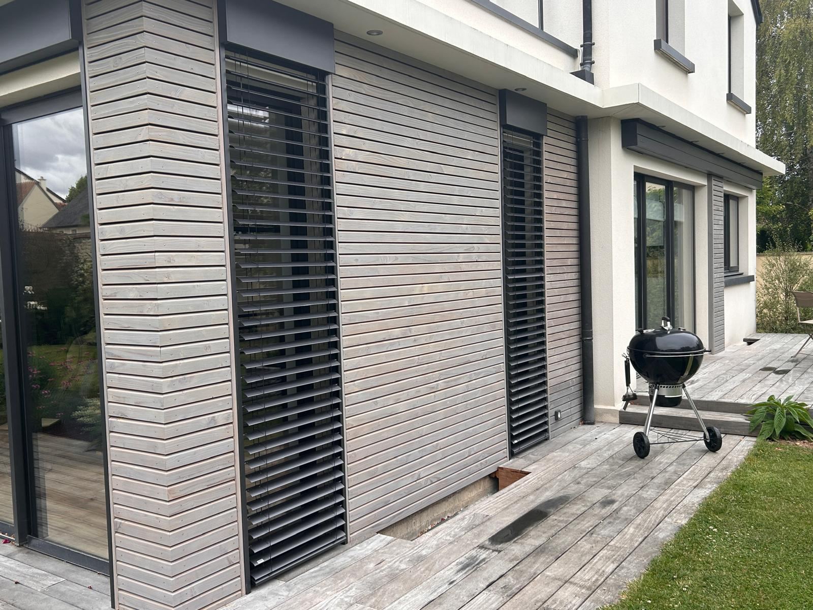 Façade bardage bois clair avec deux BSO noirs sur baies verticales et grande baie coulissante