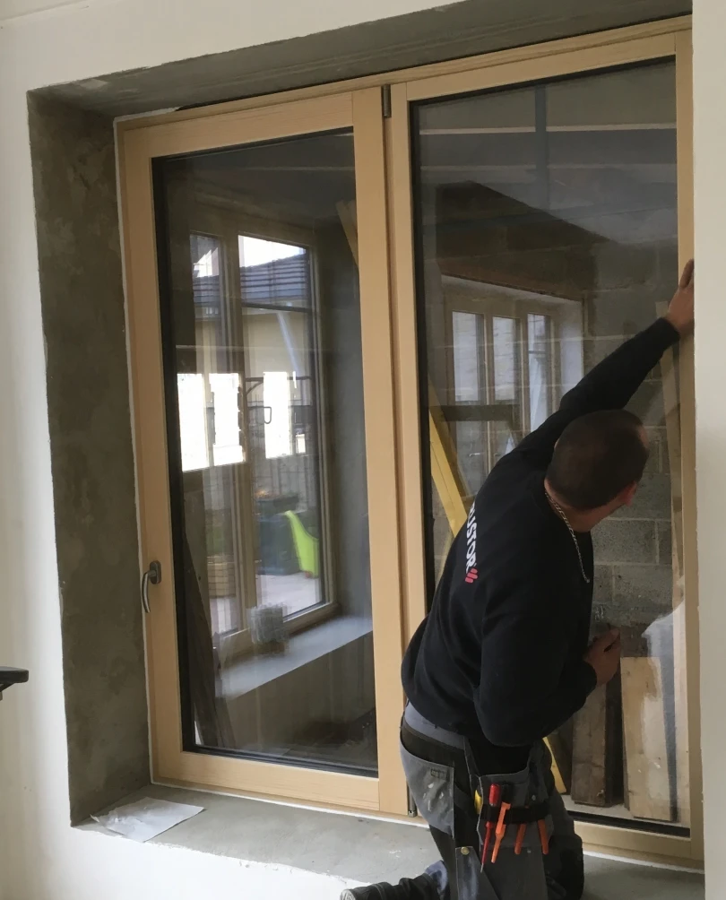 Pose fenêtre double battant bois clair par artisan, mur gris brut de chantier