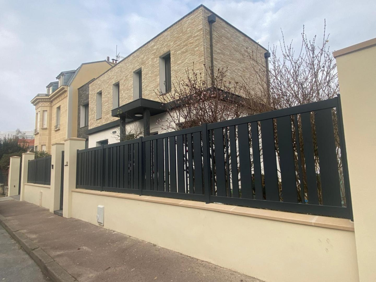 Cloture noire lames verticales sur muret beige devant maison parement pierre