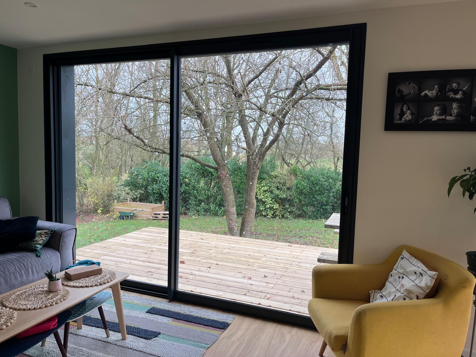 Salon avec fauteuil jaune vu à travers une baie coulissante alu noir vers terrasse bois et jardin