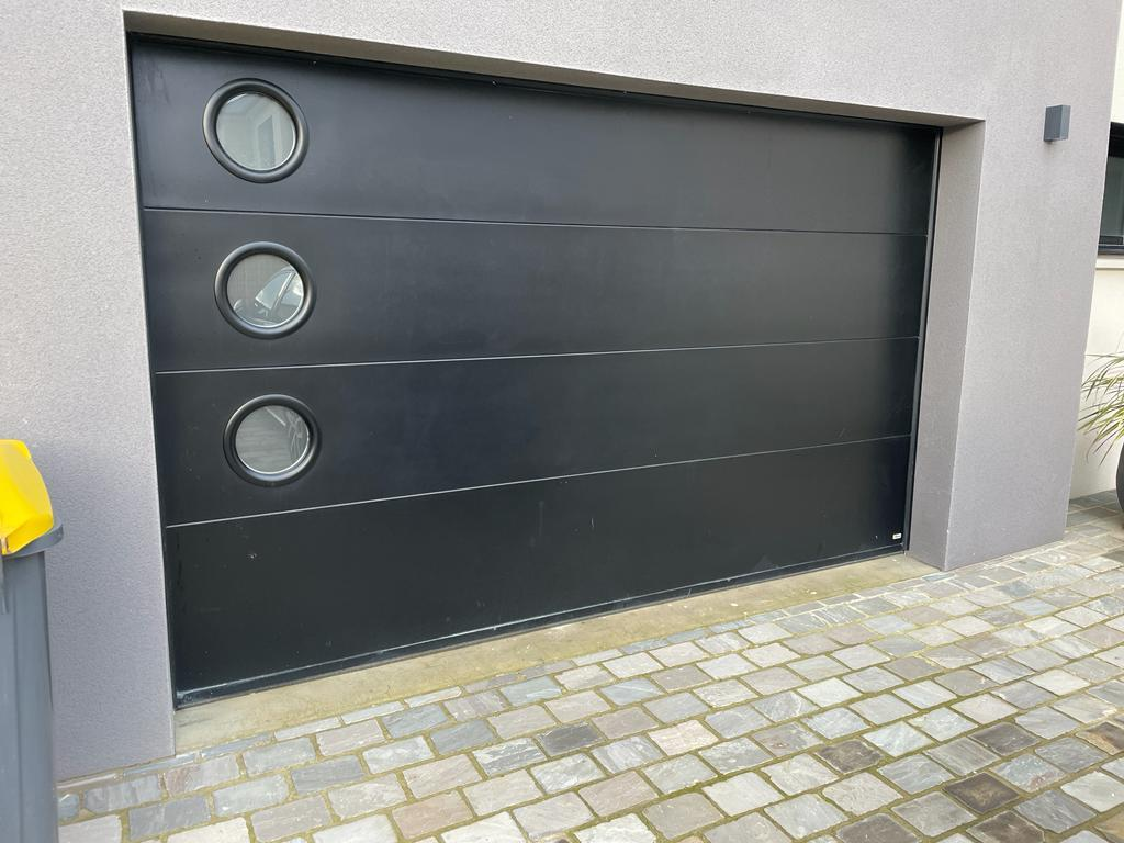 Porte garage sectionnelle noire, quatre panneaux, trois hublots ronds à gauche, pavés