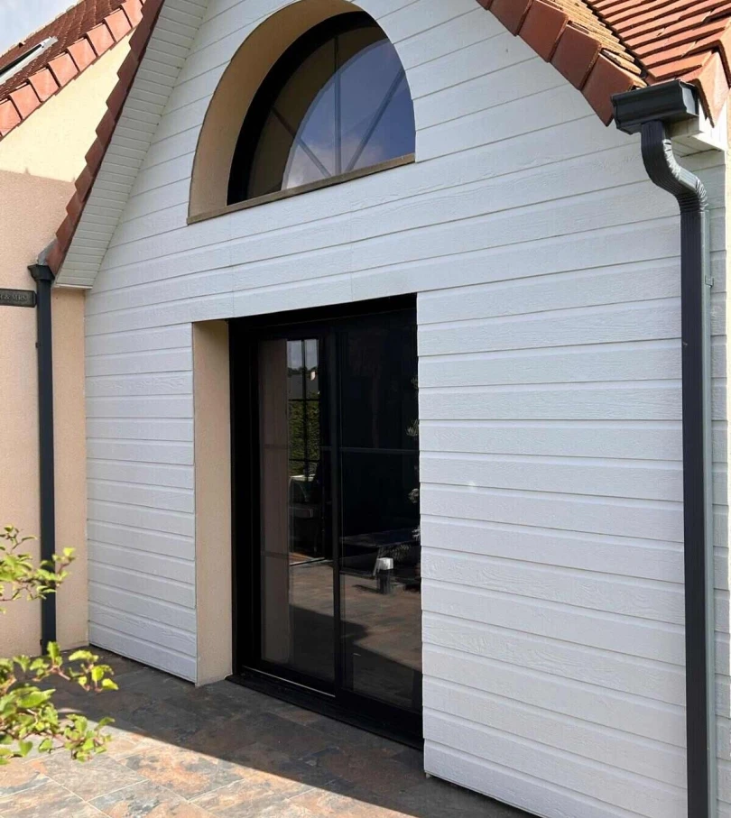 Façade bardage blanc, porte coulissante noire et fenêtre cintrée noire sous tuiles rouges