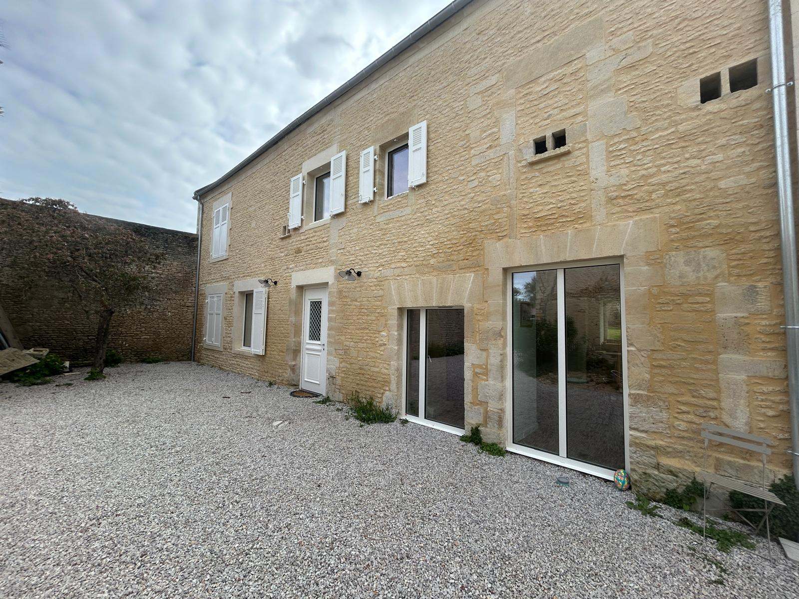 Maison en pierre rénovée avec grandes baies vitrées blanches et volets blancs, cour en gravier