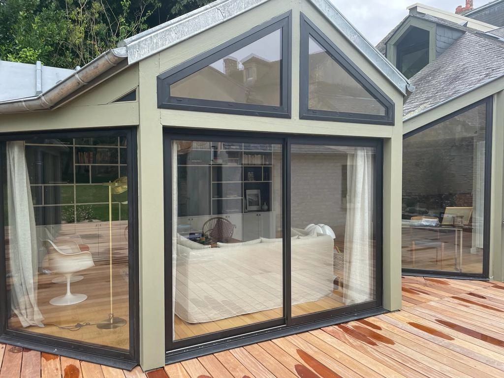 Extension vitrée vert sauge, baie coulissante marron, terrasse bois humide et bibliothèque