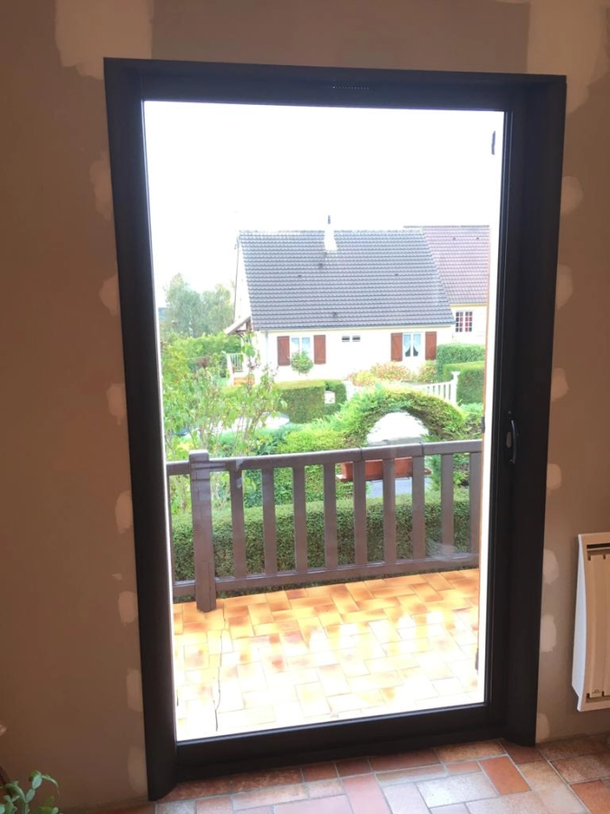 Porte-fenêtre vitrée cadre sombre, mur en rénovation, vue terrasse carrelée et jardin