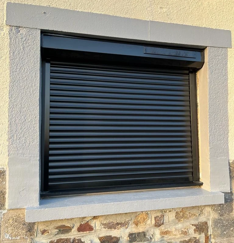 Volet roulant noir solaire fermé, panneau PV sur coffre, encadrement gris, mur pierre