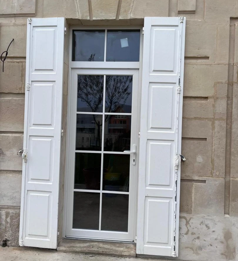 Porte vitrée blanche à croisillons, imposte et volets blancs ouverts sur mur pierre