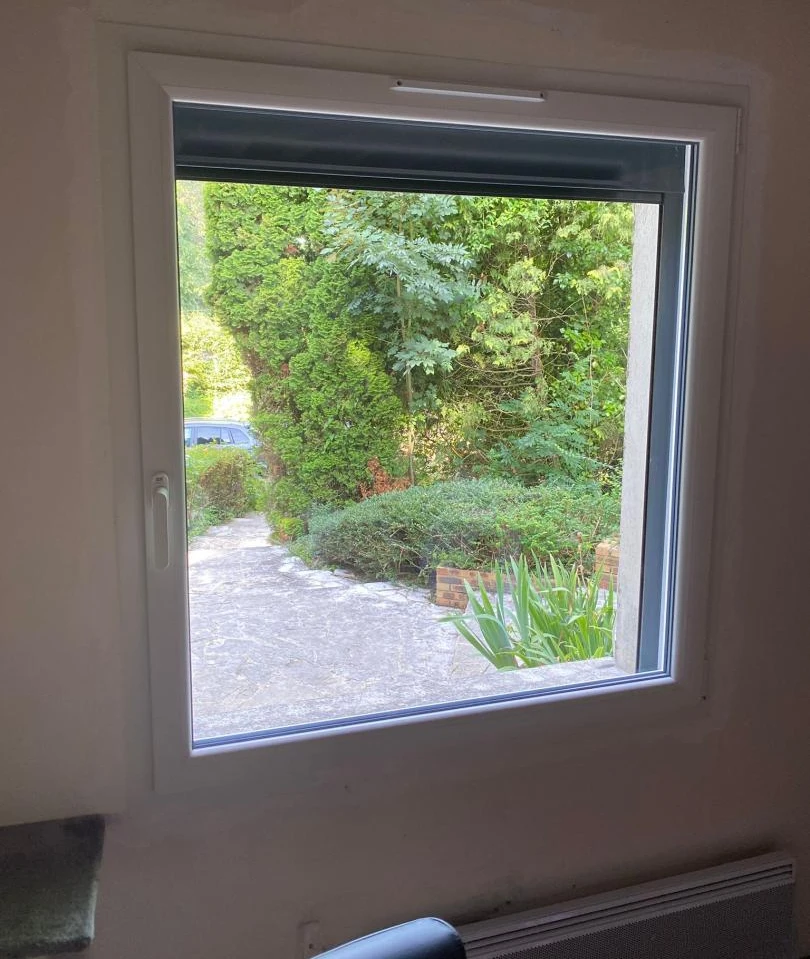 Fenêtre blanche un vantail, grille aération haute, radiateur bas et jardin ensoleillé