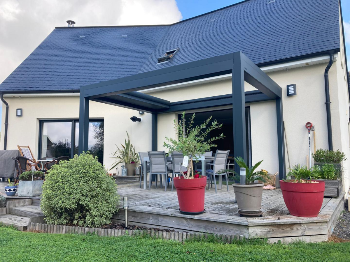 Pergola anthracite sur terrasse bois avec table grise et pots rouges devant la maison