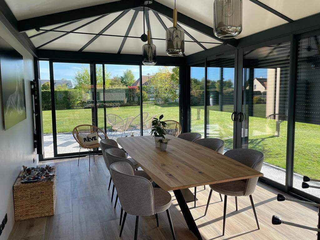 Intérieur véranda, table chêne et chaises grises, verrière noire et vue jardin ensoleillé