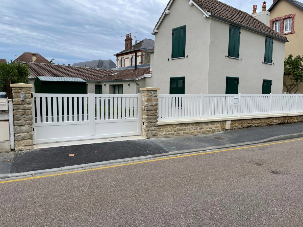 Portail blanc barreaux verticaux semi-ajouré sur muret pierre devant maison crème
