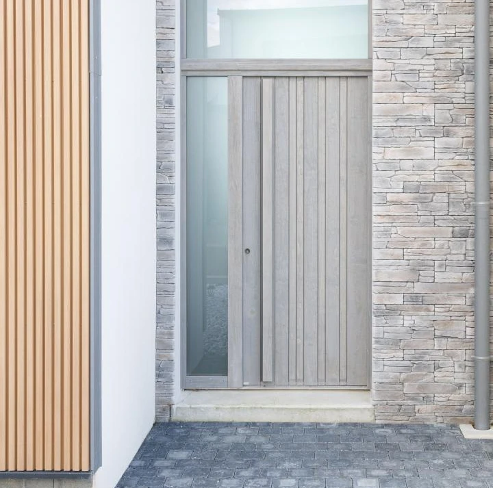 Porte d'entrée gris clair à lames verticales, tierce vitrée et imposte, mur pierre et bardage bois