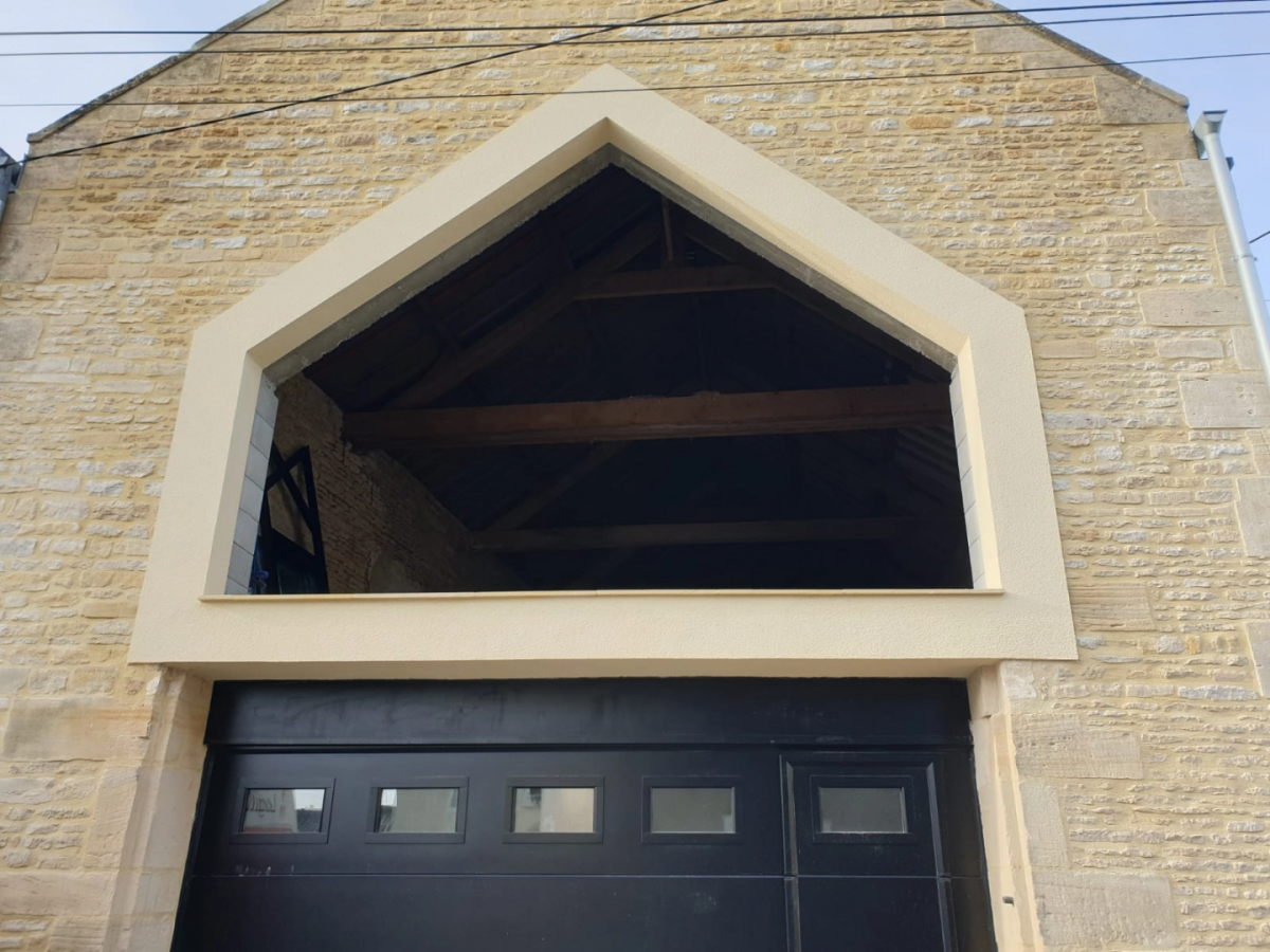 Ouverture pignon encadrée beige au-dessus porte garage noire, charpente bois visible