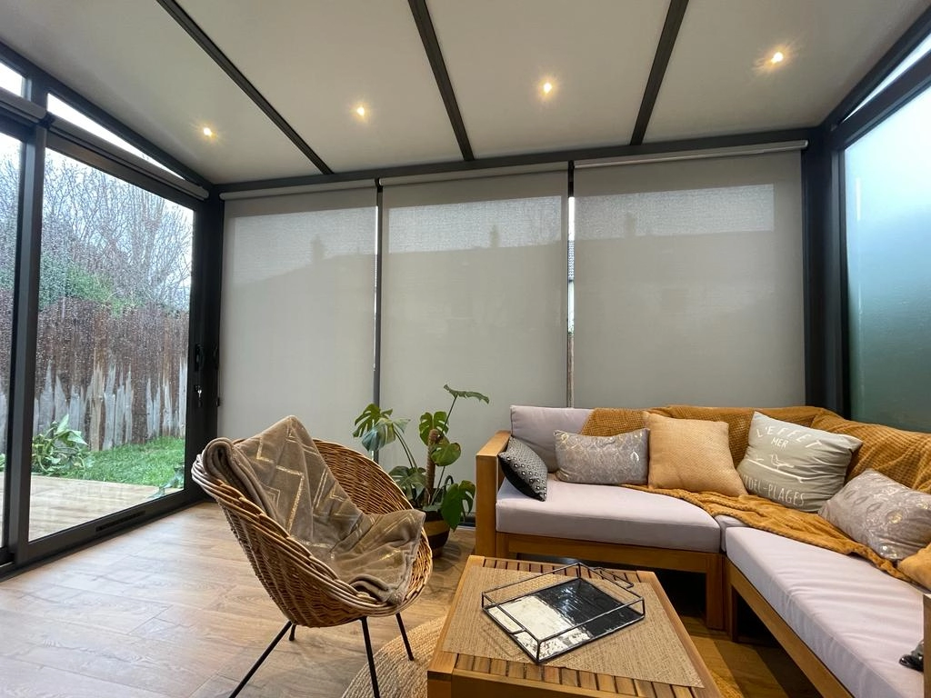 Véranda, stores enrouleurs gris, canapé angle bois, Monstera, plafond vitré éclairé