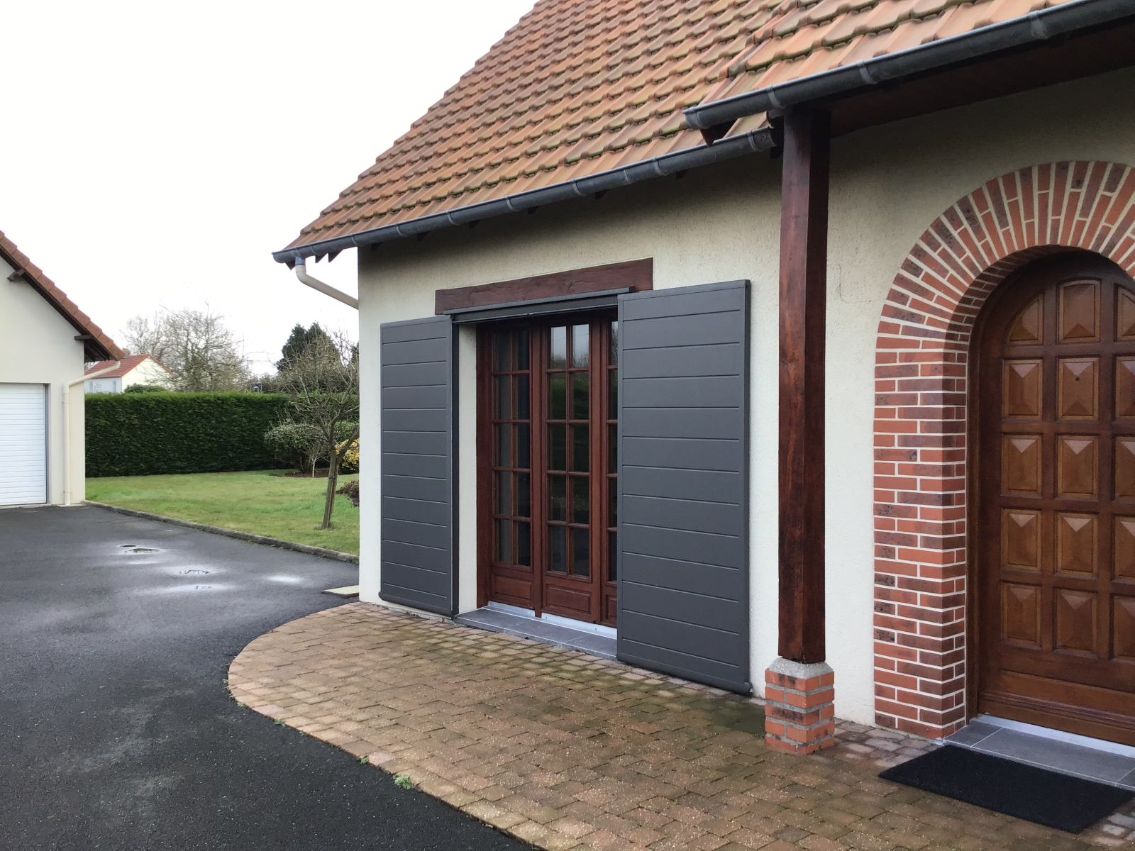 Volets coulissants anthracite à lames encadrant une porte-fenêtre bois, façade tuiles et arc brique