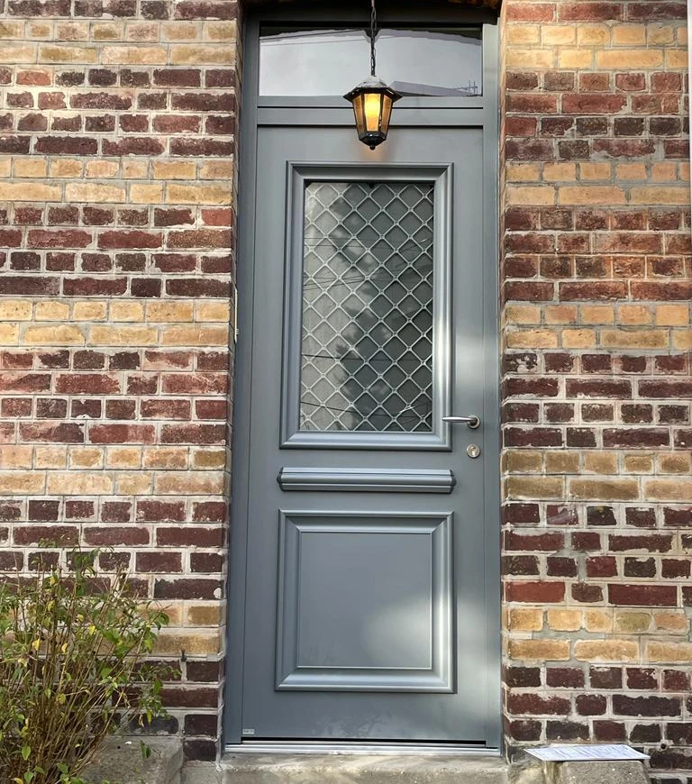 Porte gris ardoise, imposte vitrée à croisillons, lanterne noire allumée, mur brique