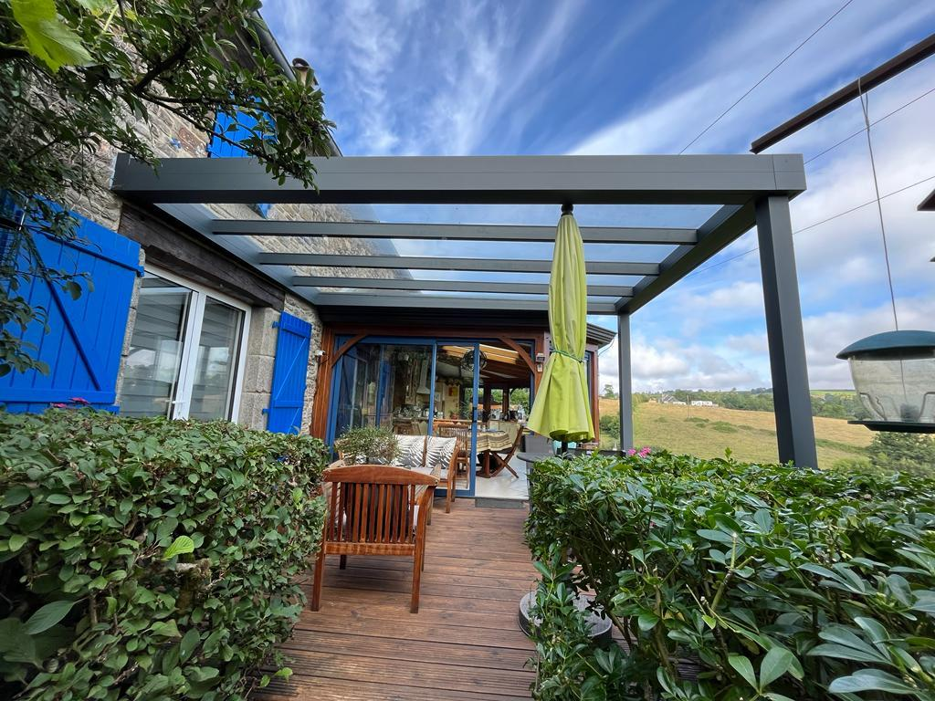 Pergola alu anthracite toit vitre sur terrasse bois, volets bleus et vue campagne