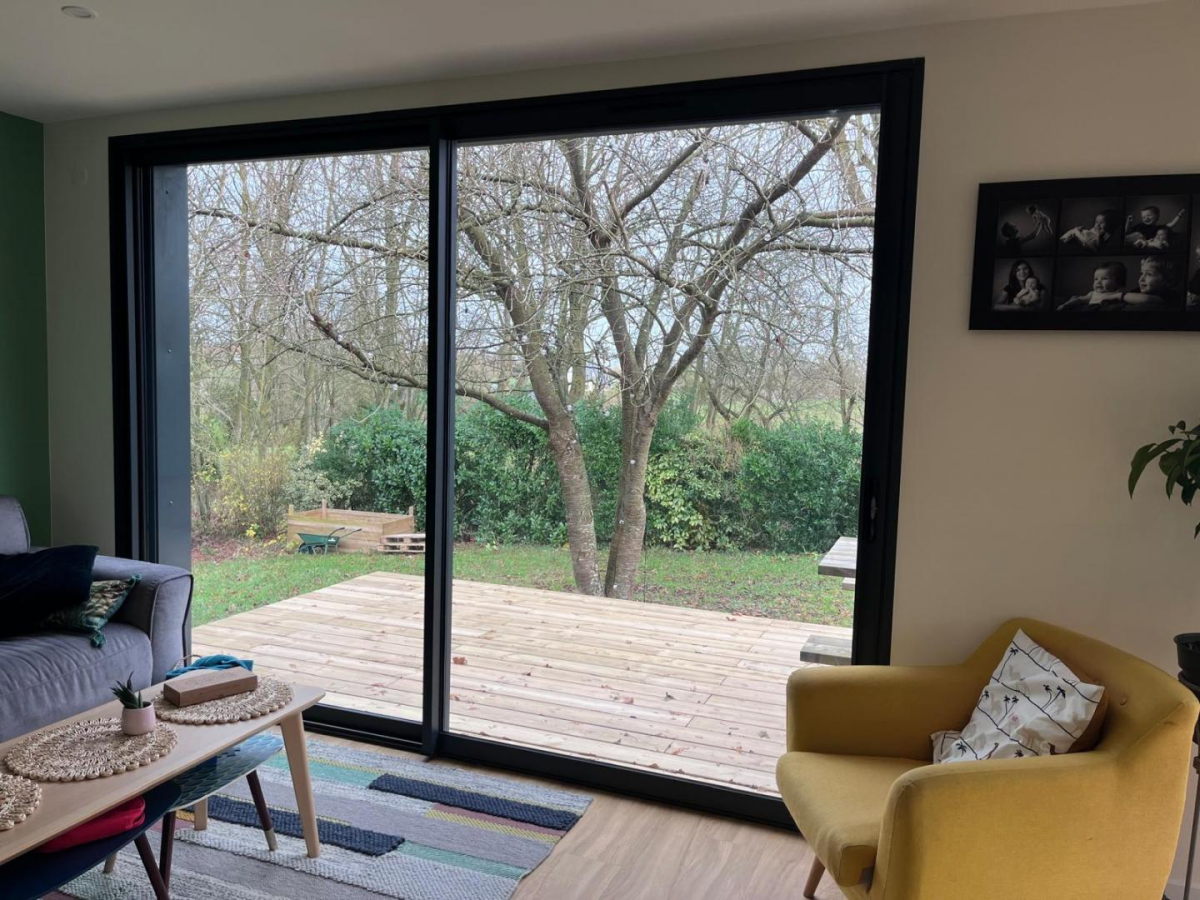 Baie coulissante noire dans salon, fauteuil jaune, parquet clair et terrasse bois jardin