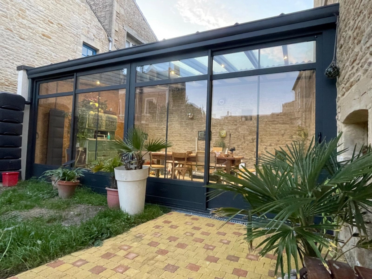 Véranda gris anthracite sur maison pierre, intérieur table bois et poêle vert