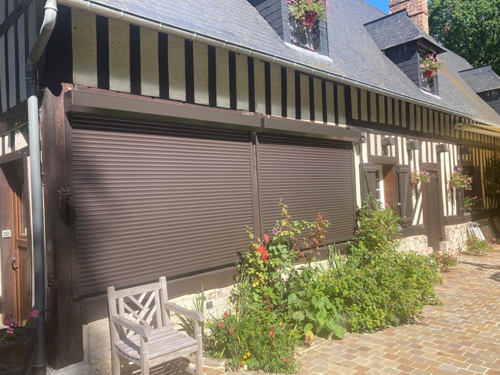 Deux volets roulants marron fermés, façade colombages, jardinières roses et chaises bois