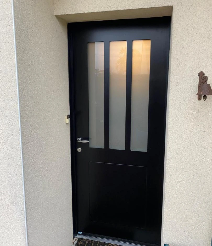 Porte d'entrée noire mate, trois vitrages verticaux dépolis, poignée chrome, mur crème