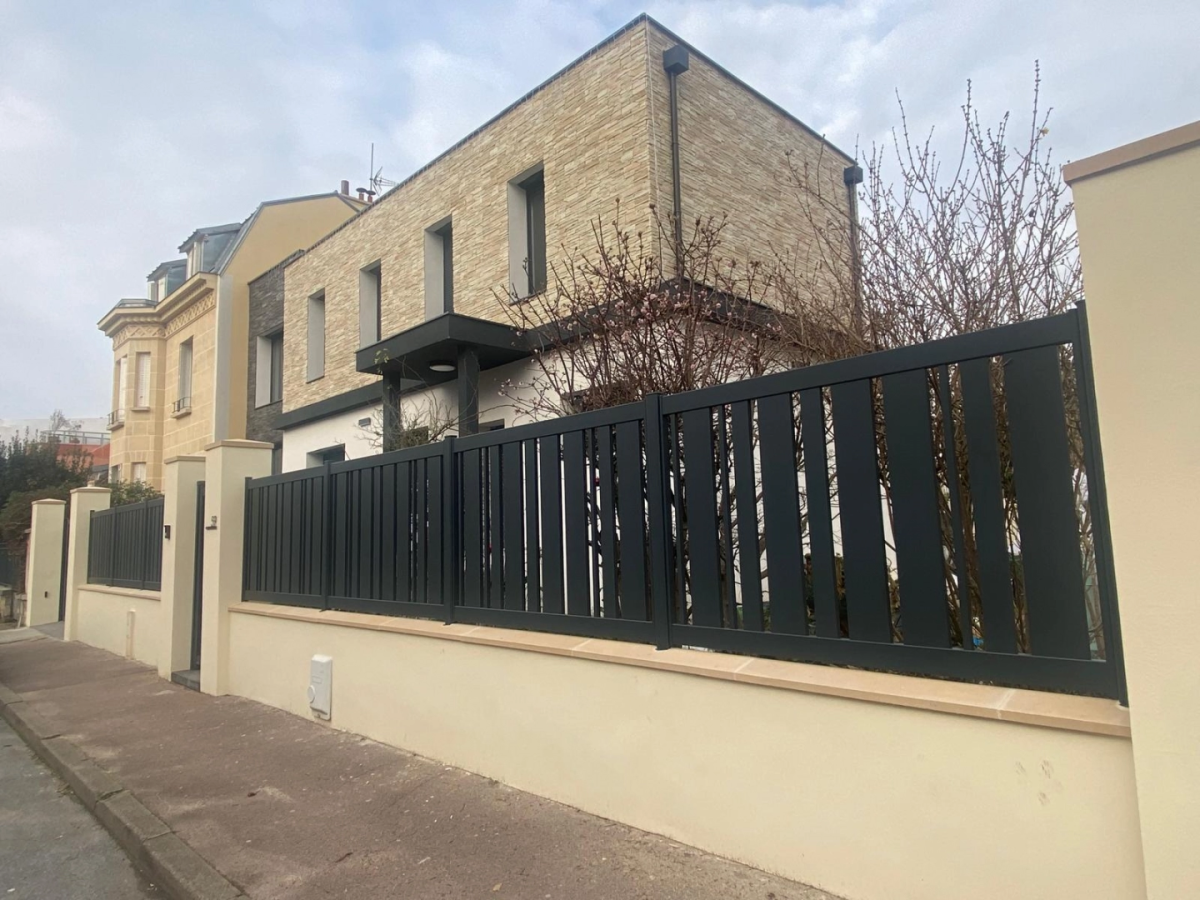 Cloture noire lames verticales alternées sur muret crème devant maison parement pierre